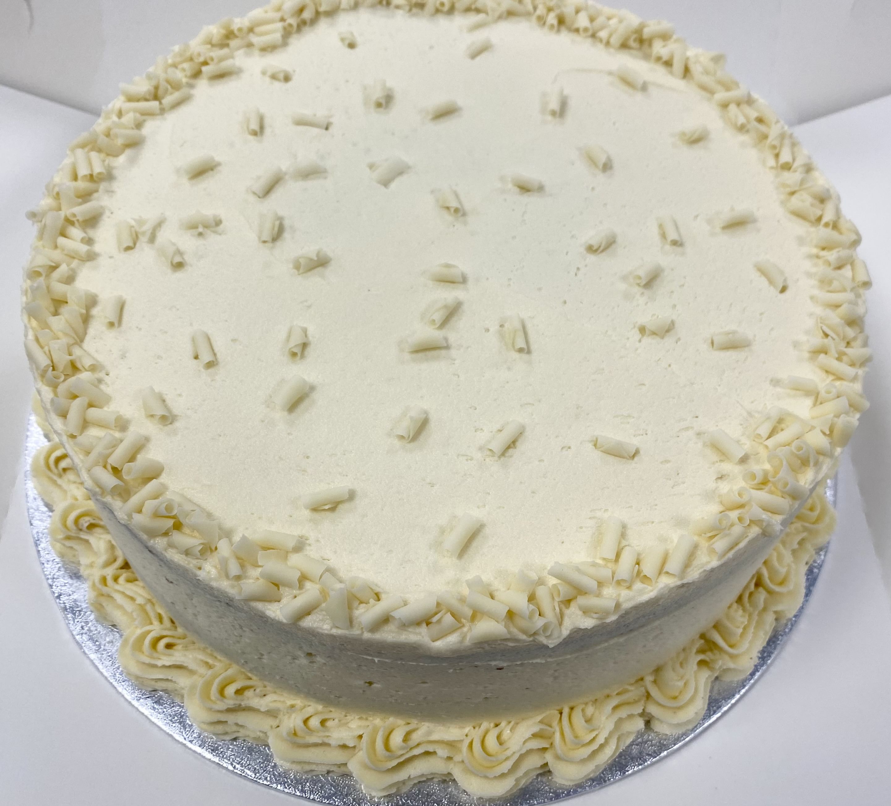 A round cake covered in smooth white frosting, topped with white chocolate shavings. It has a decorative border with a piped frosting pattern around the base. The cake is set on a silver cake board.